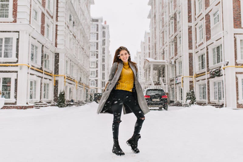 Le portrait intégral de la femme gracieuse utilise le chandail jaune à la mode dupant autour sur la rue sous des chutes de neige Photo extérieure de fille européenne mince dans la danse chaude de manteau sur le fond urbain