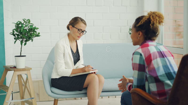 Le psychologue féminin sûr tient la consultation avec la fille d'Afro-américain, écoute la sa et écrit tandis que banque de vidéos