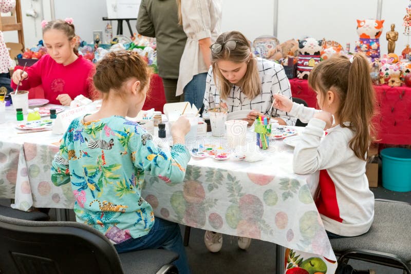 Le ragazze disegnano sotto la guida di un insegnante alla tavola nella stanza dei bambini alla fiera tradizionale popolare delle fotografia stock libera da diritti Le ragazze disegnano sotto la guida di un insegnante alla tavola nella stanza dei bambini alla fiera tradizionale popolare delle fotografia stock libera da diritti
