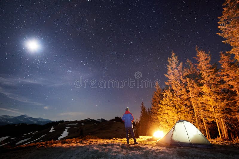 Male hiker have a rest in his camp near the forest at night. Man standing near campfire and tent under beautiful night sky full of stars and the moon, and enjoying night scene. Male hiker have a rest in his camp near the forest at night. Man standing near campfire and tent under beautiful night sky full of stars and the moon, and enjoying night scene