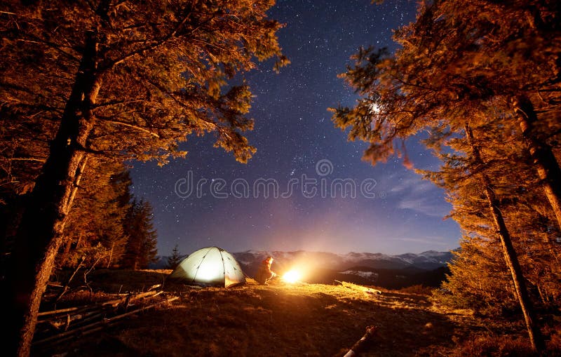 Male hiker have a rest in his camp near the forest at night. Man sitting near campfire and tent under beautiful night sky full of stars and the moon and enjoying night scene. Male hiker have a rest in his camp near the forest at night. Man sitting near campfire and tent under beautiful night sky full of stars and the moon and enjoying night scene