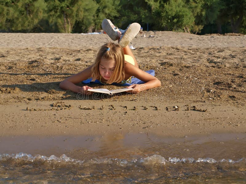 A view at a child - young girl female lying and reading a open book at sandy beach in summer vacation. Horizontal color photo. A view at a child - young girl female lying and reading a open book at sandy beach in summer vacation. Horizontal color photo.