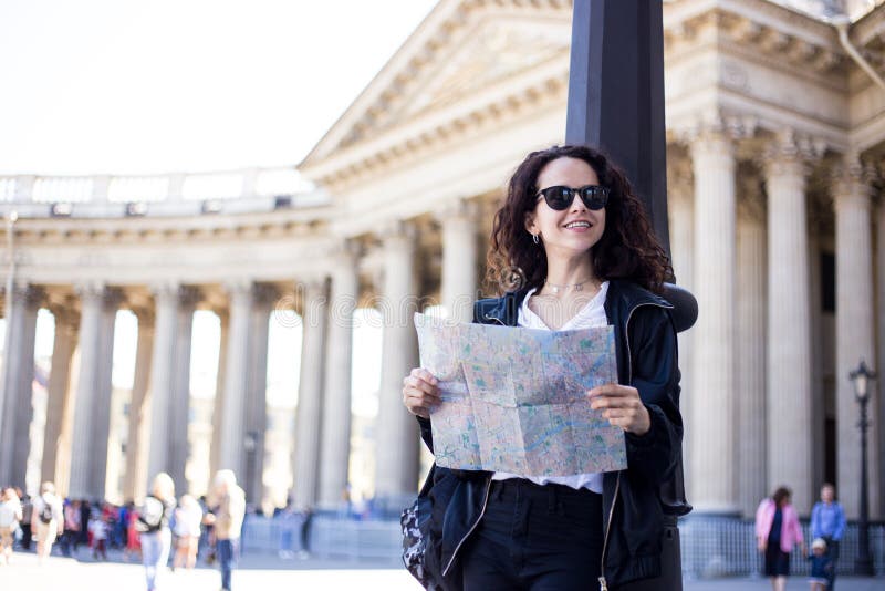 Le sac à dos de port de sourire de femme de voyageur et les lunettes de soleil noires tiennent la carte de site dans des mains, p photographie stock libre de droits Le sac à dos de port de sourire de femme de voyageur et les lunettes de soleil noires tiennent la carte de site dans des mains, p photographie stock libre de droits