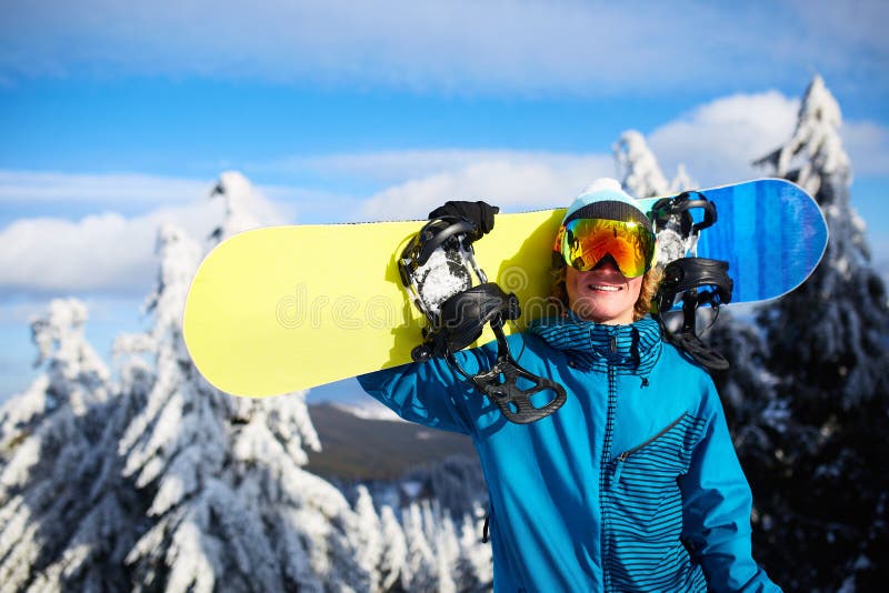 Le snowboarderen som poserar den bärande snowboarden på skuldra på, skida semesterorten nära skog för backcountry freeride och arkivbild Le snowboarderen som poserar den bärande snowboarden på skuldra på, skida semesterorten nära skog för backcountry freeride och arkivbild