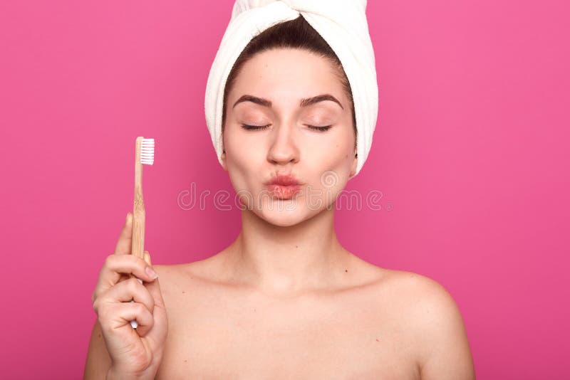Le studio tiré de la femelle heureuse avec des yeux fermés, boude des lèvres, porte la serviette blanche sur la tête, brosse à de image stock Le studio tiré de la femelle heureuse avec des yeux fermés, boude des lèvres, porte la serviette blanche sur la tête, brosse à de image stock