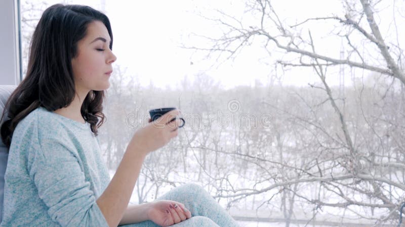 Le temps à la maison, la belle femme avec la tasse de café dans des mains rêve près de la fenêtre dans la chambre