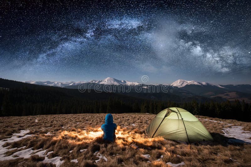 Female tourist have a rest in her camp at night. Woman sitting near campfire and green tent under beautiful sky full of stars and milky way. On the background snow-covered mountains. Long exposure. Female tourist have a rest in her camp at night. Woman sitting near campfire and green tent under beautiful sky full of stars and milky way. On the background snow-covered mountains. Long exposure