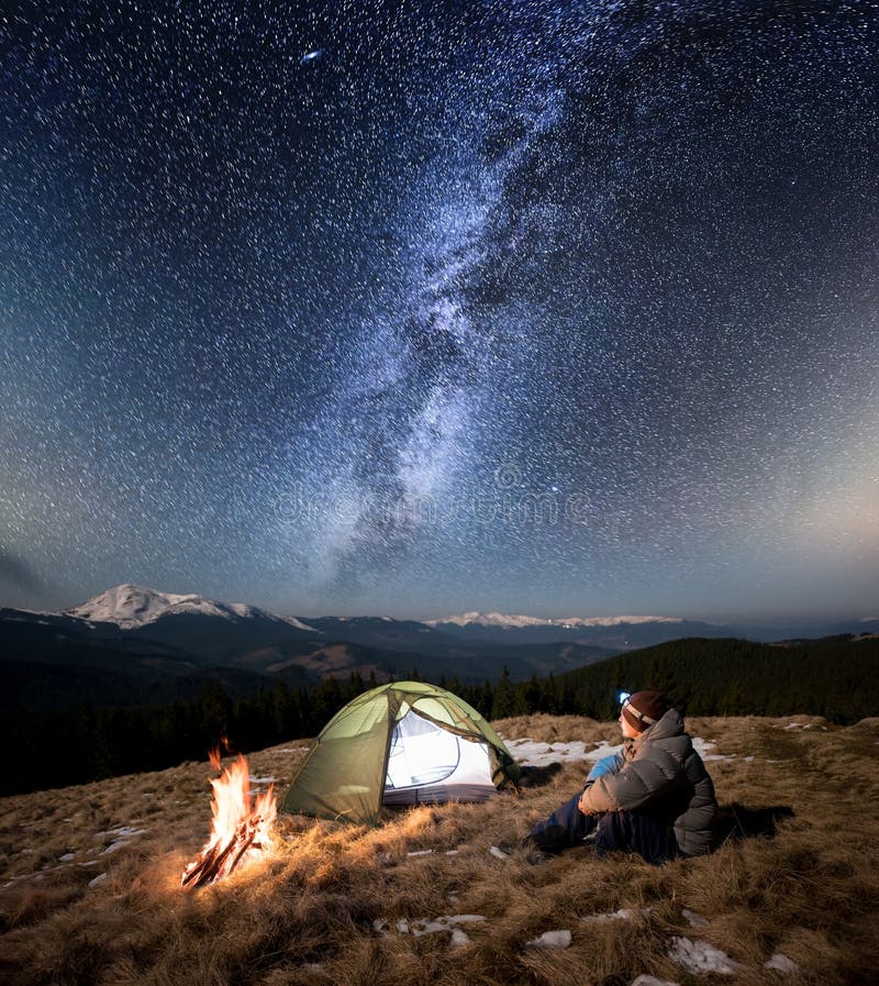 Male tourist have a rest in his camp at night. Man with a headlamp sitting near campfire and tent under beautiful sky full of stars and milky way. On the background snow-covered mountains and forests. Male tourist have a rest in his camp at night. Man with a headlamp sitting near campfire and tent under beautiful sky full of stars and milky way. On the background snow-covered mountains and forests