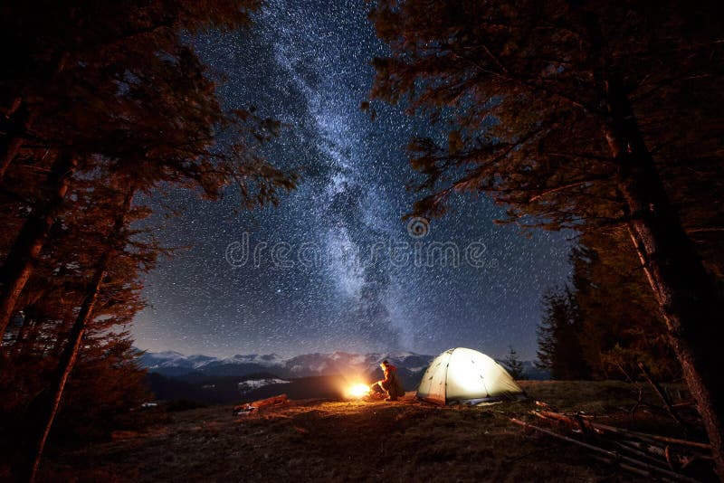 Male tourist have a rest in his camp near the forest at night. Man sitting near campfire and tent under beautiful night sky full of stars and milky way, and enjoying night scene. Astrophotography. Male tourist have a rest in his camp near the forest at night. Man sitting near campfire and tent under beautiful night sky full of stars and milky way, and enjoying night scene. Astrophotography