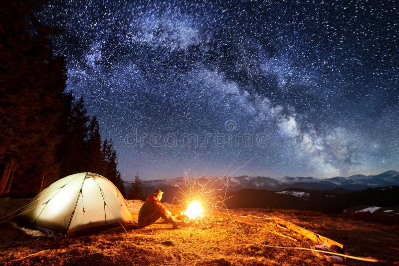 Male tourist have a rest in his camp near the forest at night. Man sitting near campfire and tent under beautiful night sky full of stars and milky way, and enjoying night scene. Male tourist have a rest in his camp near the forest at night. Man sitting near campfire and tent under beautiful night sky full of stars and milky way, and enjoying night scene