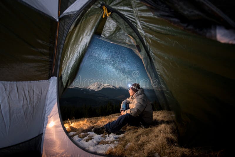 View from inside a tent on the male tourist have a rest in his camping in the mountains at night. Man with a headlamp sitting near campfire under beautiful night sky full of stars and milky way. View from inside a tent on the male tourist have a rest in his camping in the mountains at night. Man with a headlamp sitting near campfire under beautiful night sky full of stars and milky way