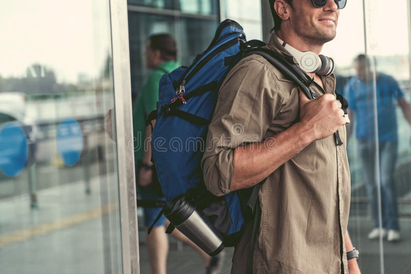 Le type agréable positif avec des écouteurs sort du bâtiment d'aéroport. Jour merveilleux Le jeune homme mignon avec le sac à dos et les lunettes de soleil est extinction des portes Il exprime le bonheur Copiez l'espace dans le côté gauche