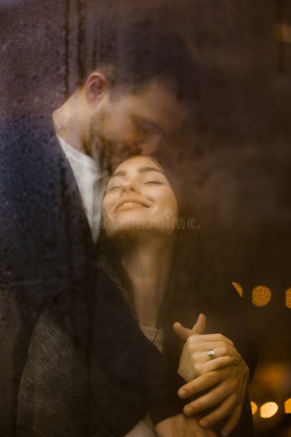 Le type aimant étreint et embrasse sa position heureuse d'amie derrière une fenêtre humide avec des lumières Couples romantiques photo libre de droits Le type aimant étreint et embrasse sa position heureuse d'amie derrière une fenêtre humide avec des lumières Couples romantiques photo libre de droits