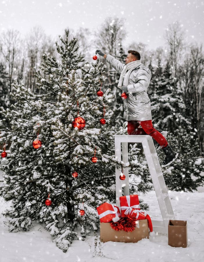 Le type de jeune homme dans des vêtements d'hiver élégant décore l'arbre de Noël avec des balles en plein air dans la forêt neigeu
