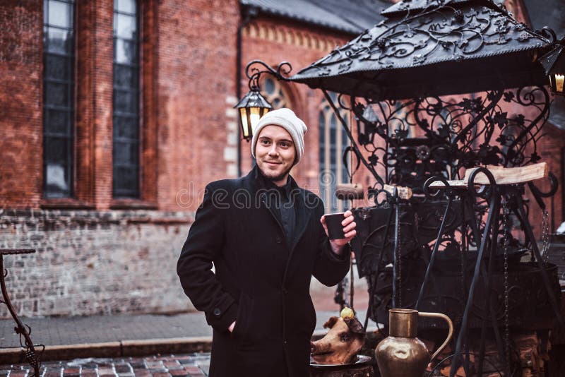 Le type gai tient une tasse de café et de regards en longueur tout en marchant par la foire de Noël photographie stock Le type gai tient une tasse de café et de regards en longueur tout en marchant par la foire de Noël photographie stock