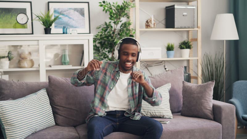 Le type heureux d'Afro-américain est dansant et chantant écouter la musique par des écouteurs se reposant sur le divan à la maiso banque de vidéos