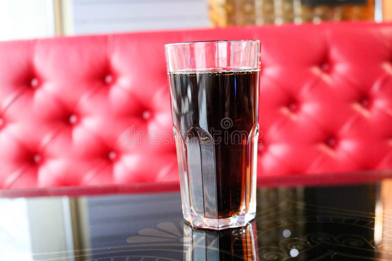 Le verre avec un brun, bonbon, froid, a carbonaté la boisson sur une table dans un café le soir sur le fond d'un sofa rouge photographie stock Le verre avec un brun, bonbon, froid, a carbonaté la boisson sur une table dans un café le soir sur le fond d'un sofa rouge photographie stock