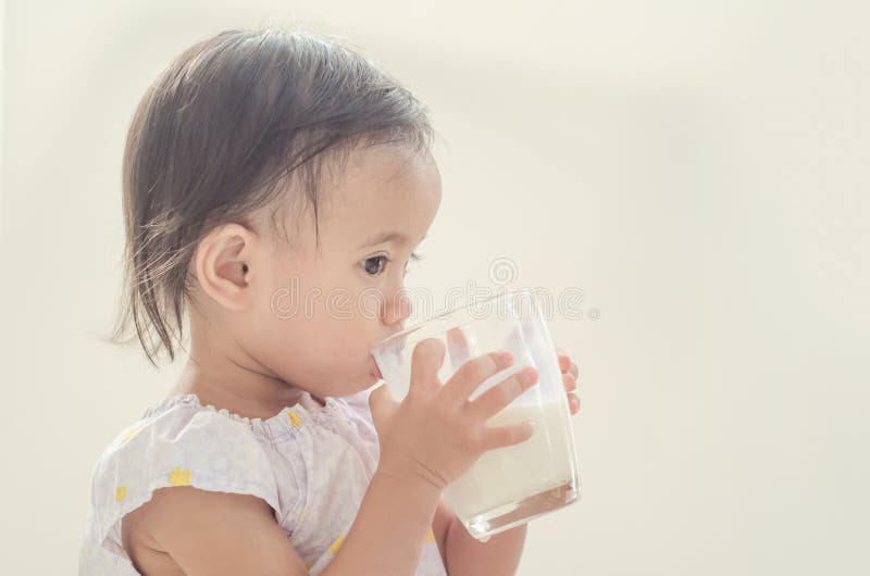 Leche de consumo de la niña pequeña asiática linda del vidrio grande en los vagos blancos imagenes de archivo Leche de consumo de la niña pequeña asiática linda del vidrio grande en los vagos blancos imagenes de archivo