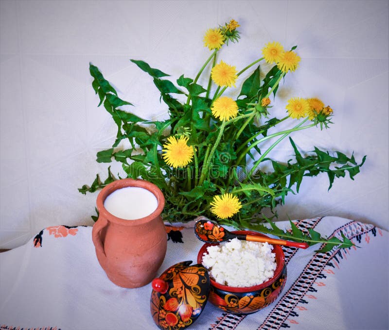 Leche y queso cottege sobre la mesa. En el mantel bordado hay flores de león y leche con queso de cabaña