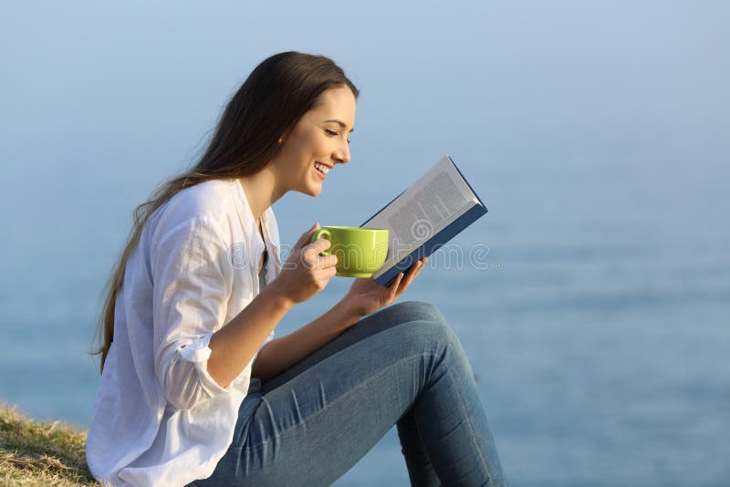 Side view portrait of a happy girl relaxing reading a book and drinking coffee sitting on the grass on the beach. Side view portrait of a happy girl relaxing reading a book and drinking coffee sitting on the grass on the beach