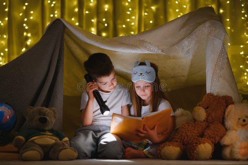 Lecturi și jocuri de familie în cortul copiilor. băiat și fată cu carte și lanternă înainte de a merge la culcare stock image