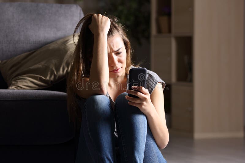 Front view of a sad teen checking phone sitting on the floor in the living room at home with a dark background. Front view of a sad teen checking phone sitting on the floor in the living room at home with a dark background