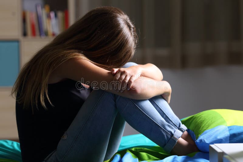 Portrait of a single sad teen sitting on the bed in her bedroom with a dark light in the background. Portrait of a single sad teen sitting on the bed in her bedroom with a dark light in the background