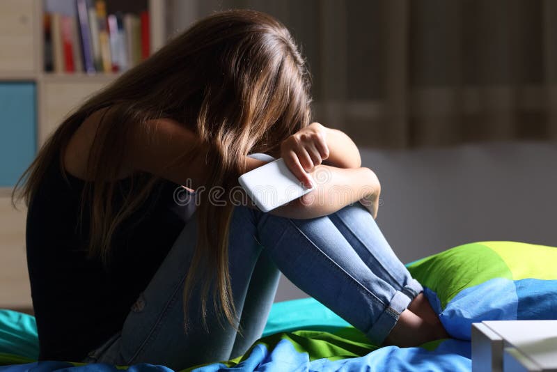 Single sad teen holding a mobile phone lamenting sitting on the bed in her bedroom with a dark light in the background. Single sad teen holding a mobile phone lamenting sitting on the bed in her bedroom with a dark light in the background