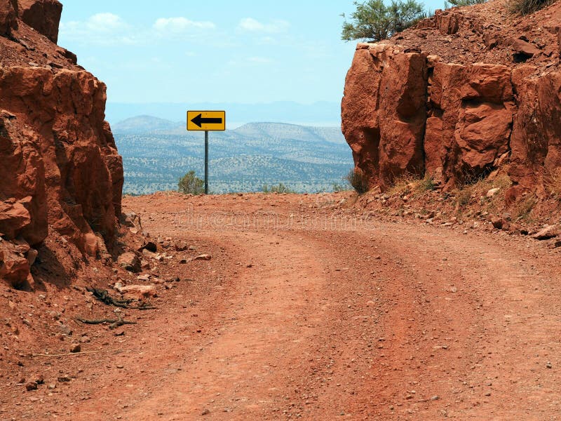 A Yellow and Black Left Arrow Sign on the dangerous curve of a mountain dirt road. A Yellow and Black Left Arrow Sign on the dangerous curve of a mountain dirt road.