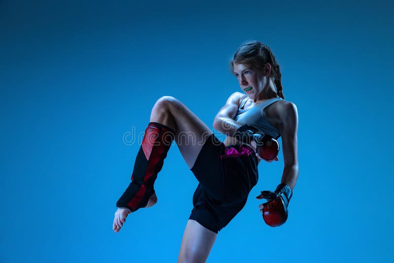 Studio shot of sportive teen-girl, MMA fighter in action, motion isolated on blue background in neon light. Concept of