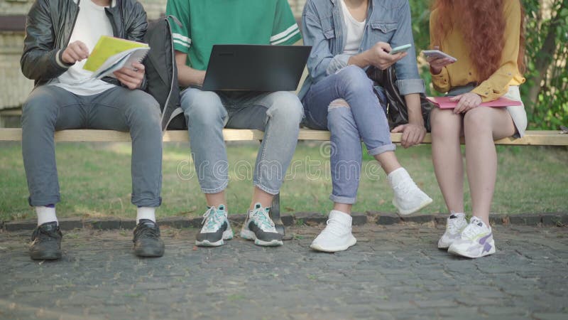 Legs of four Caucasian university students sitting on bench outdoors surfing Internet and doing homework. Unrecognizable
