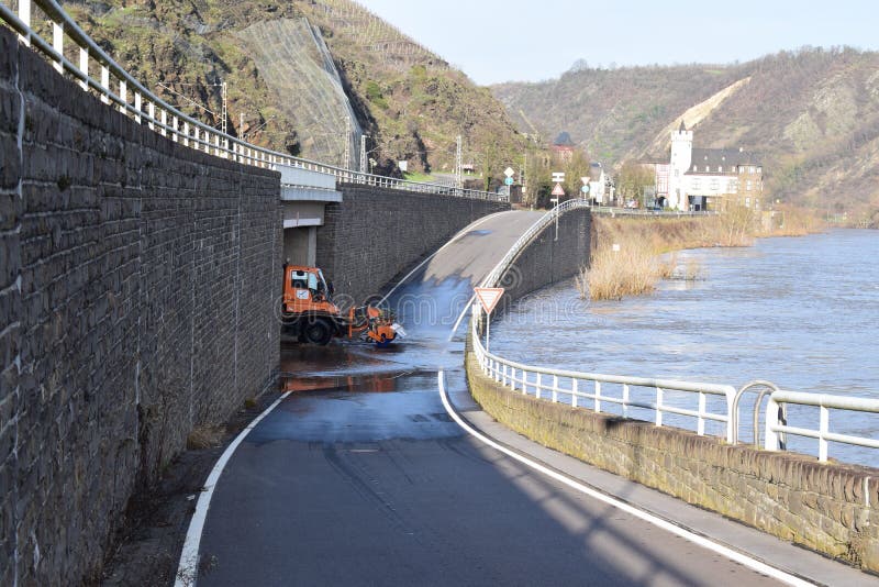 Mosel valley road near Lehmen during the winter flood with muddy water on the road intersection. A road maintenance truck is using a rotating brush on the ground to remove mud while the river Moselle is still bringing water back on the street. The brushed of muddy water goes back into the river flood, being on road level. It is a small winter flood. Mosel valley road near Lehmen during the winter flood with muddy water on the road intersection. A road maintenance truck is using a rotating brush on the ground to remove mud while the river Moselle is still bringing water back on the street. The brushed of muddy water goes back into the river flood, being on road level. It is a small winter flood