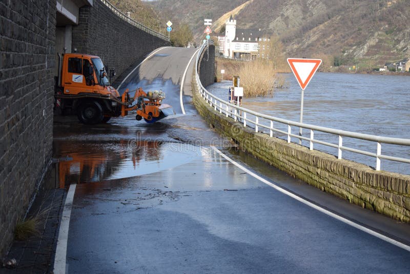 Mosel valley road near Lehmen during the winter flood with muddy water on the road intersection. A road maintenance truck is using a rotating brush on the ground to remove mud while the river Moselle is still bringing water back on the street. The brushed of muddy water goes back into the river flood, being on road level. It is a small winter flood. Mosel valley road near Lehmen during the winter flood with muddy water on the road intersection. A road maintenance truck is using a rotating brush on the ground to remove mud while the river Moselle is still bringing water back on the street. The brushed of muddy water goes back into the river flood, being on road level. It is a small winter flood