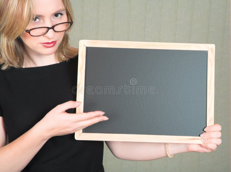 Young serious blond woman teacher in black dress and pearls, wearing glasses pointing at empty blackboard. Young serious blond woman teacher in black dress and pearls, wearing glasses pointing at empty blackboard