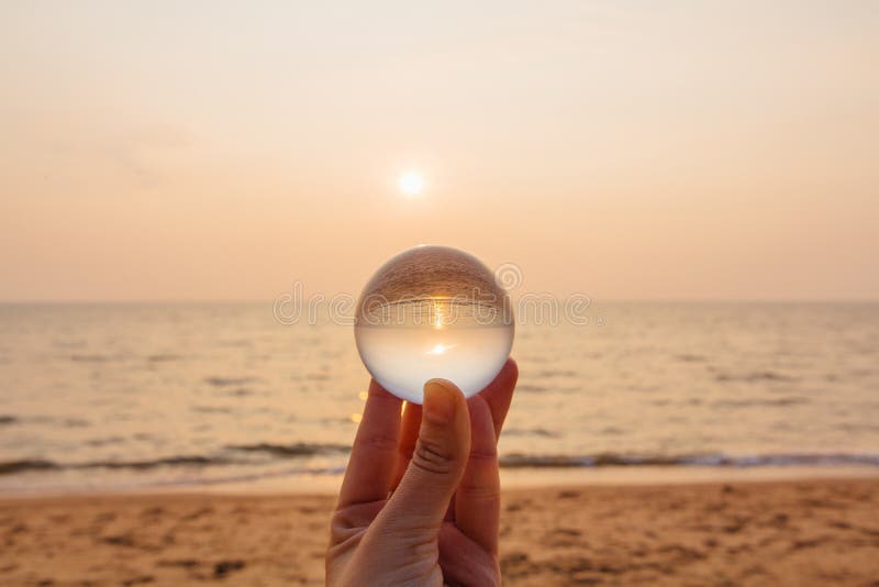 Lens boll i hand med reflexion av havet och solnedgången på stranden