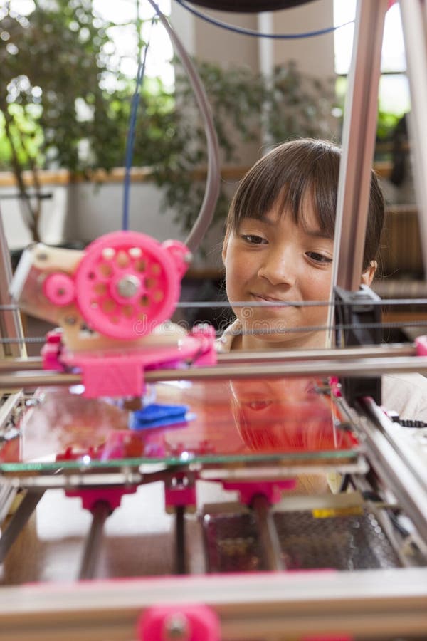 A young girl watches as a 3D printer prints an object. A young girl watches as a 3D printer prints an object.