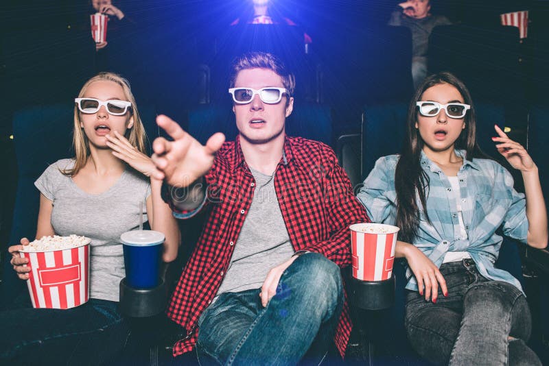 Amazed and surprised teenagers are watching 3d movie. THey wear glasses for that. Guy is trying to reach the screen with his hand. Girls are sitting besides him having popcorn. Amazed and surprised teenagers are watching 3d movie. THey wear glasses for that. Guy is trying to reach the screen with his hand. Girls are sitting besides him having popcorn