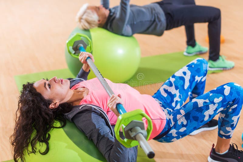 Les athlètes féminins faisant le coffre de barbell pressent le mensonge sur une boule suisse pendant la formation de groupe au ce photos stock Les athlètes féminins faisant le coffre de barbell pressent le mensonge sur une boule suisse pendant la formation de groupe au ce photos stock