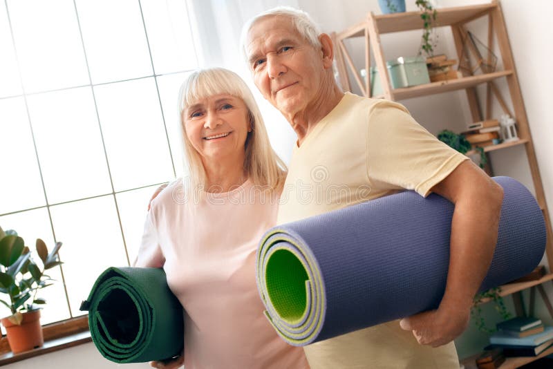 Les couples supérieurs exercent ensemble à la maison des soins de santé tenant des tapis de yoga regardant l'appareil-photo photo stock Les couples supérieurs exercent ensemble à la maison des soins de santé tenant des tapis de yoga regardant l'appareil-photo photo stock