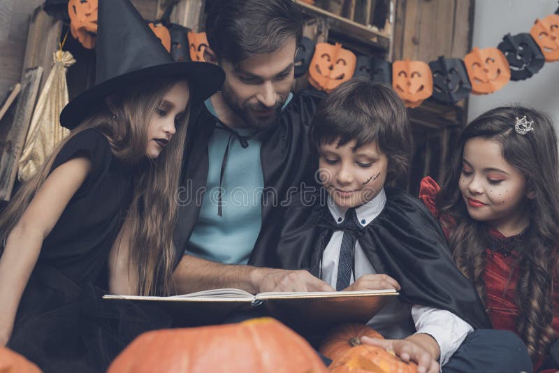 Les enfants dans des costumes de monstre s'asseyent autour d'un homme dans un manteau noir et lisent un livre à une partie de Hal photos stock Les enfants dans des costumes de monstre s'asseyent autour d'un homme dans un manteau noir et lisent un livre à une partie de Hal photos stock