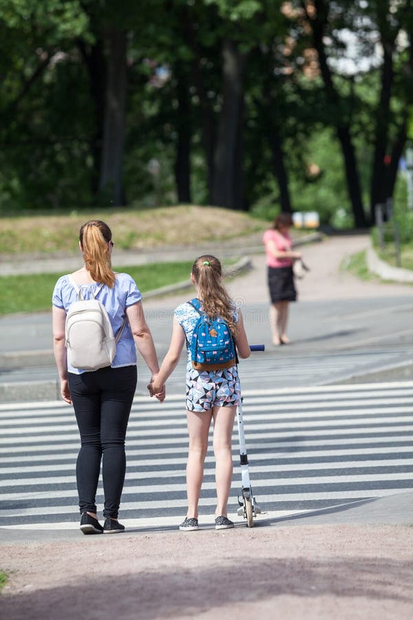 People standing at urban crosswalk, child with kick scooter hold mothers hand, red light. Rear view. People standing at urban crosswalk, child with kick scooter hold mothers hand, red light. Rear view