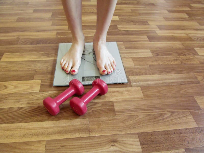Les jambes femelles se tiennent sur les échelles et le mensonge rouge de deux haltères près sur le plancher photographie stock Les jambes femelles se tiennent sur les échelles et le mensonge rouge de deux haltères près sur le plancher photographie stock