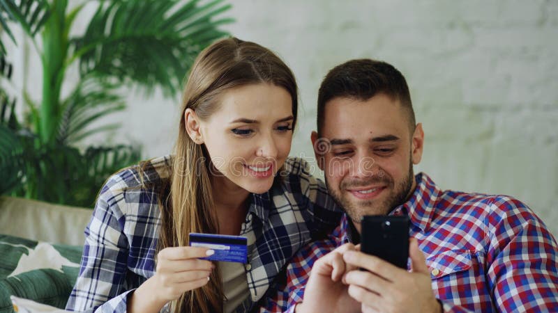 Les jeunes ajouter attrayants au smartphone et les achats de carte de crédit sur l'Internet se reposent sur le divan dans le salo photos stock Les jeunes ajouter attrayants au smartphone et les achats de carte de crédit sur l'Internet se reposent sur le divan dans le salo photos stock