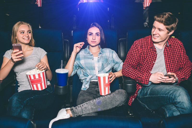 Very good young friends are sitting together in cinema. Blonde gir and boy are looking at each other and smiling. She is holding phone in hands. Brunette is looking straight and eating popcorn. Very good young friends are sitting together in cinema. Blonde gir and boy are looking at each other and smiling. She is holding phone in hands. Brunette is looking straight and eating popcorn