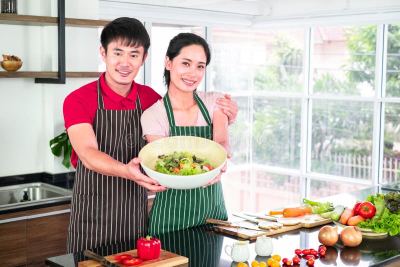 Les jeunes couples asiatiques sourient, se réjouissent à l'apron, préparent ensemble leur cuisine Montrer un grand bol de salade photo libre de droits Les jeunes couples asiatiques sourient, se réjouissent à l'apron, préparent ensemble leur cuisine Montrer un grand bol de salade photo libre de droits