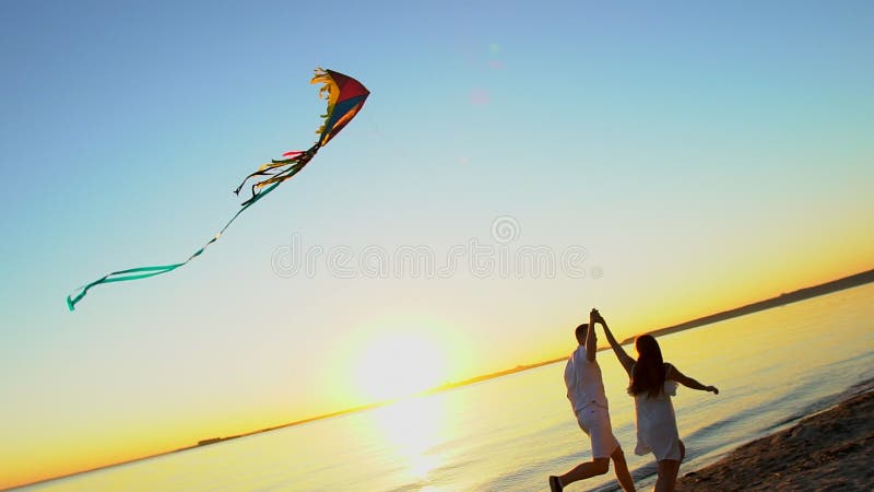 Les jeunes couples heureux jouant avec un cerf-volant sur la mer échouent au coucher du soleil Homme et femme appréciant le temps banque de vidéos