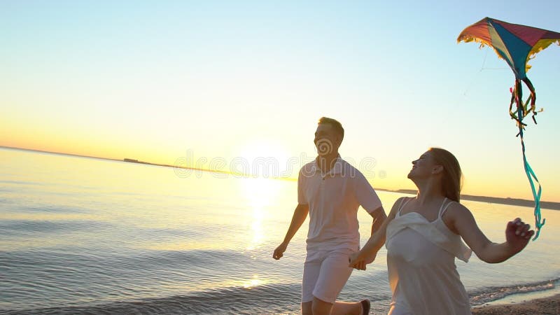 Les jeunes couples heureux jouant avec un cerf-volant sur la mer échouent au coucher du soleil Homme et femme appréciant le temps banque de vidéos