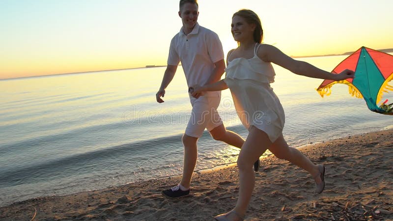 Les jeunes couples heureux jouant avec un cerf-volant sur la mer échouent au coucher du soleil Homme et femme appréciant le temps banque de vidéos