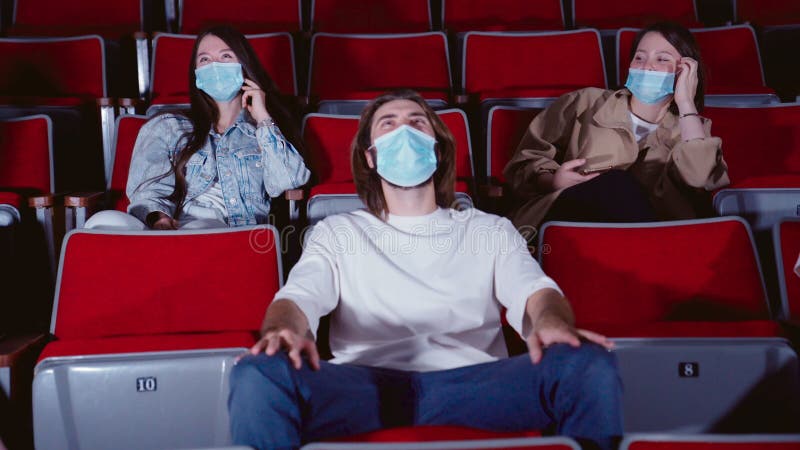 Young peoplein medical masks laughing at cinema theater. Joyful Cheerful audience watching amusing comedy in cinema and wearing medical masks, prevention of covid 19 virus. Young peoplein medical masks laughing at cinema theater. Joyful Cheerful audience watching amusing comedy in cinema and wearing medical masks, prevention of covid 19 virus.