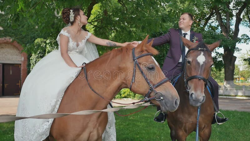 Les jeunes mariés, tenant des mains, s'asseyent sur les chevaux magnifiques en beau parc vert le jour de leur mariage